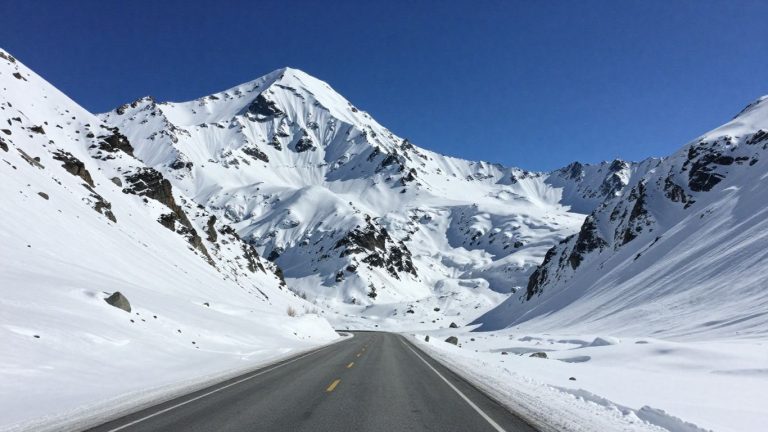 Snowy Alaskan highway amidst majestic mountains under blue sky.